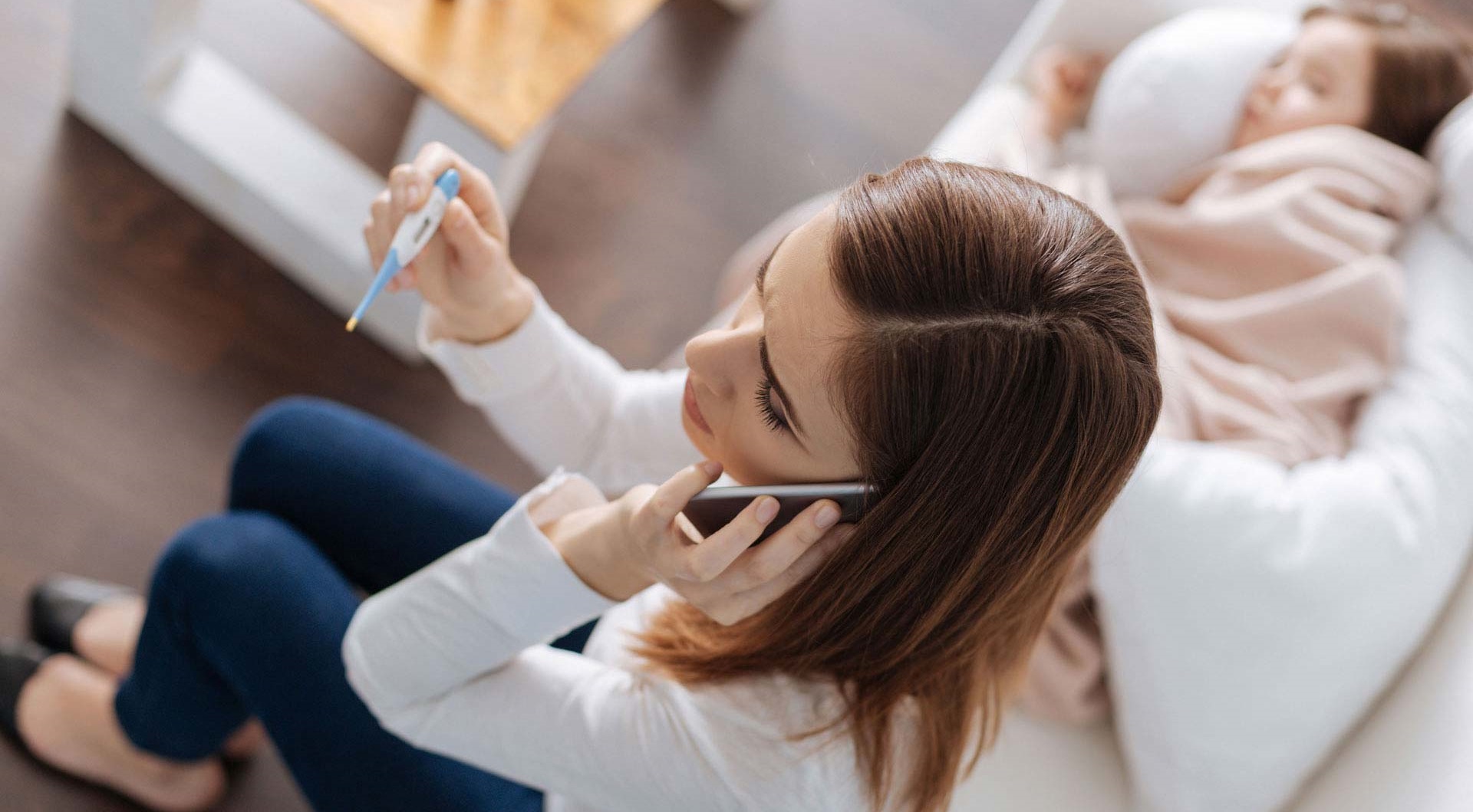 Frau hält Fieberthermometer in der einen Hand und mit der anderen Hand hält sie sich das Telefon ans Ohr.